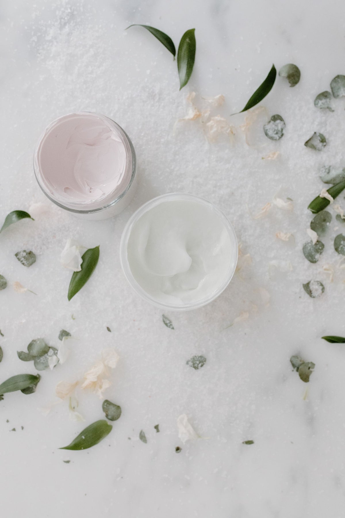 Zwei geöffnete Cremetiegel auf Marmor, umgeben von Blättern und Blüten